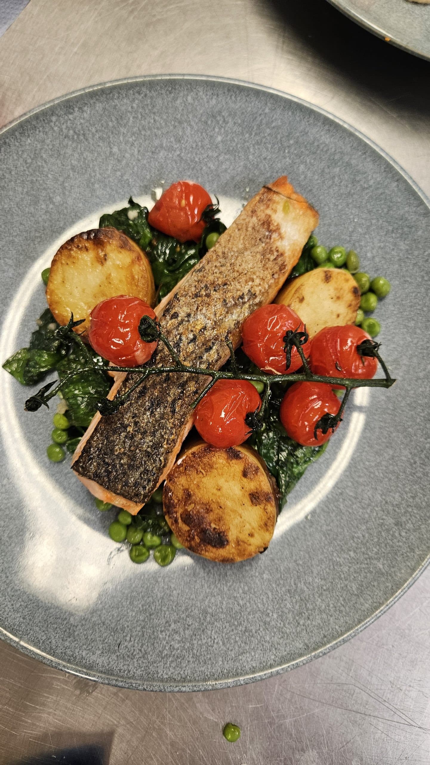 A plated fine dining dish featuring a seared fish fillet with herb crust, accompanied by roasted tomatoes, sautéed spinach, seared potato rounds, and fresh green peas on a grey ceramic plate