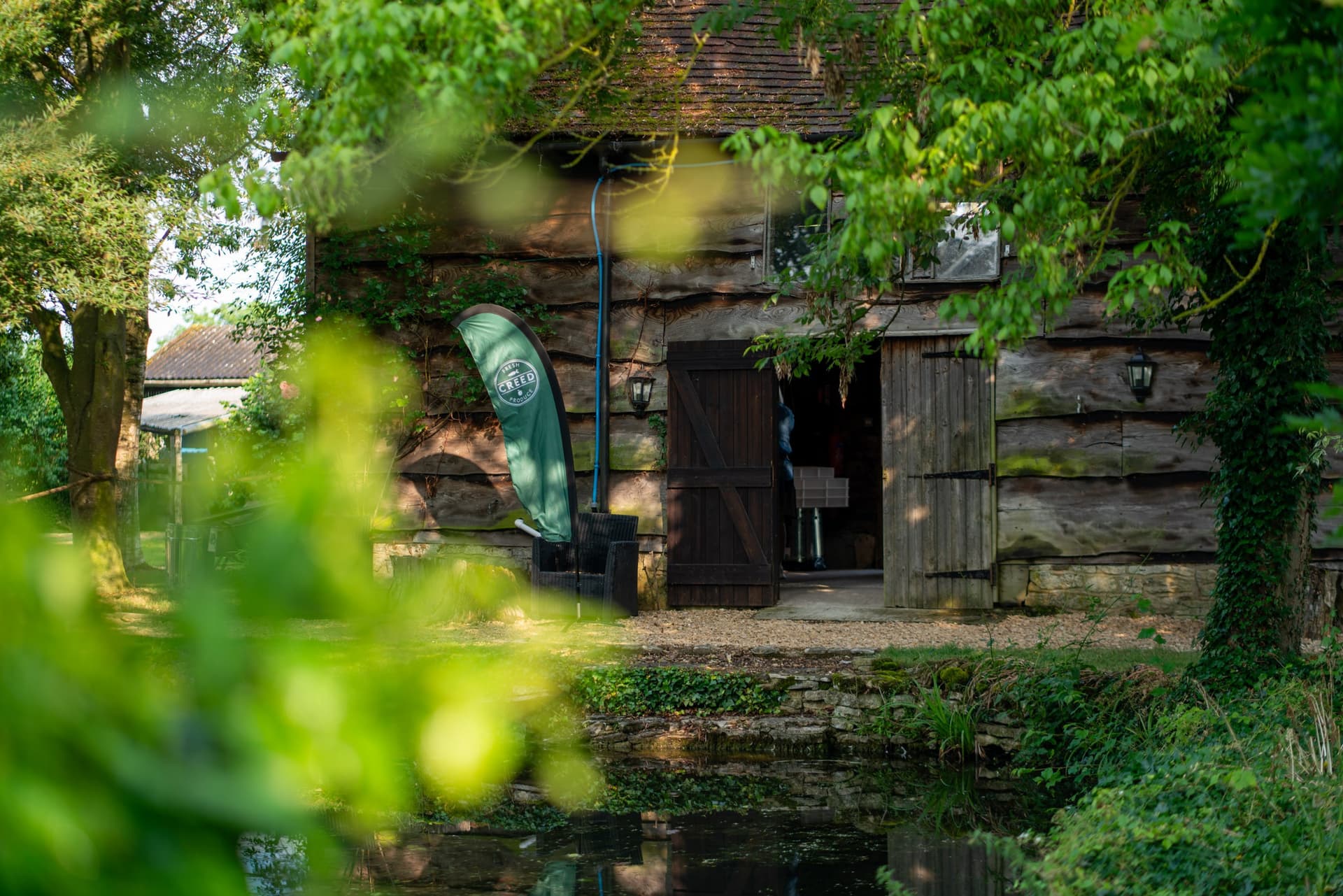 A rustic stone building with ivy-covered walls, timber doors, and a green teepee-shaped banner at the entrance, framed by lush green foliage and vegetation