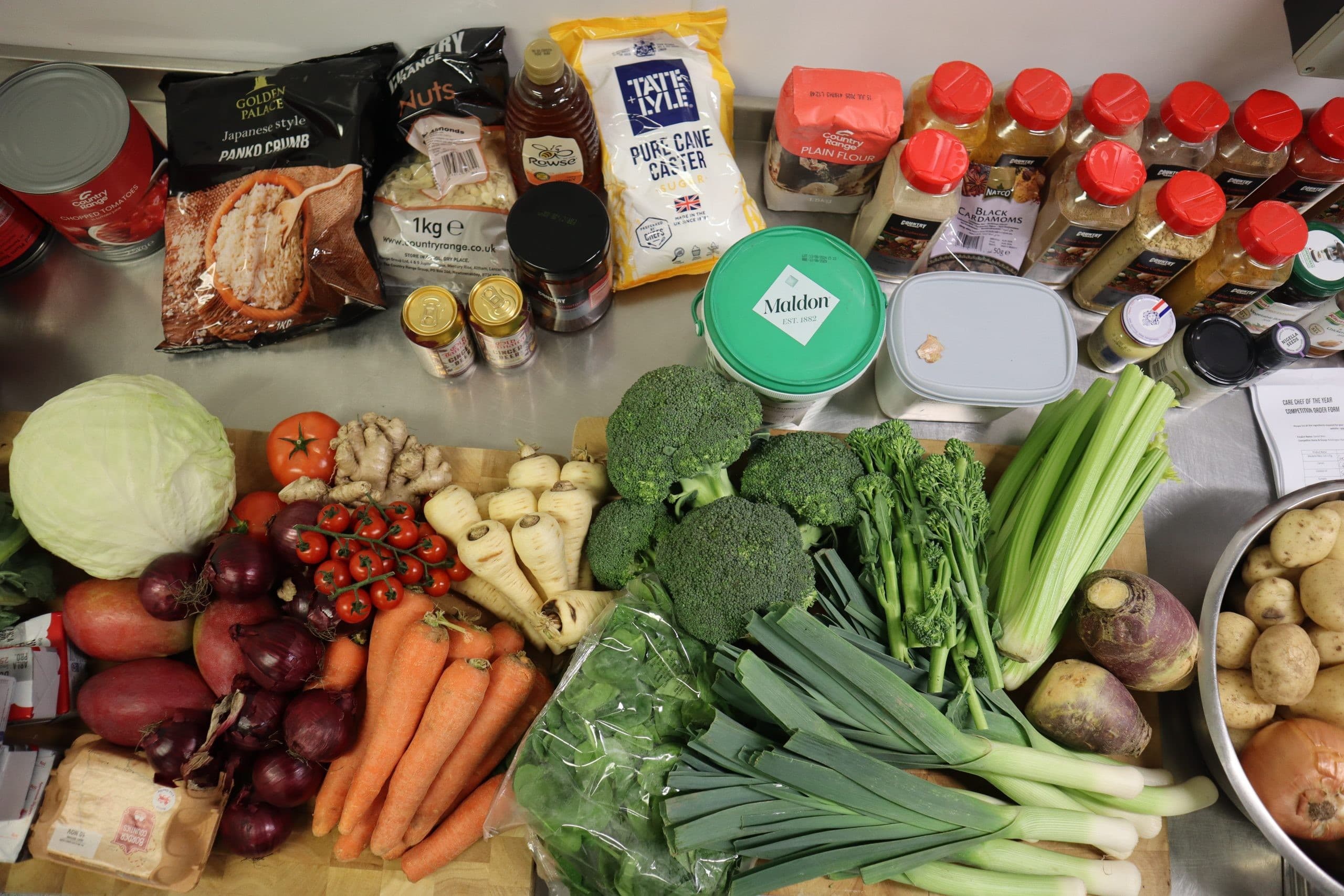 A flat lay photograph of fresh vegetables, packaged ingredients, and condiments arranged on a grey work surface, including broccoli, leeks, carrots, onions, tomatoes, cabbage, and various packaged foods with jars and containers.