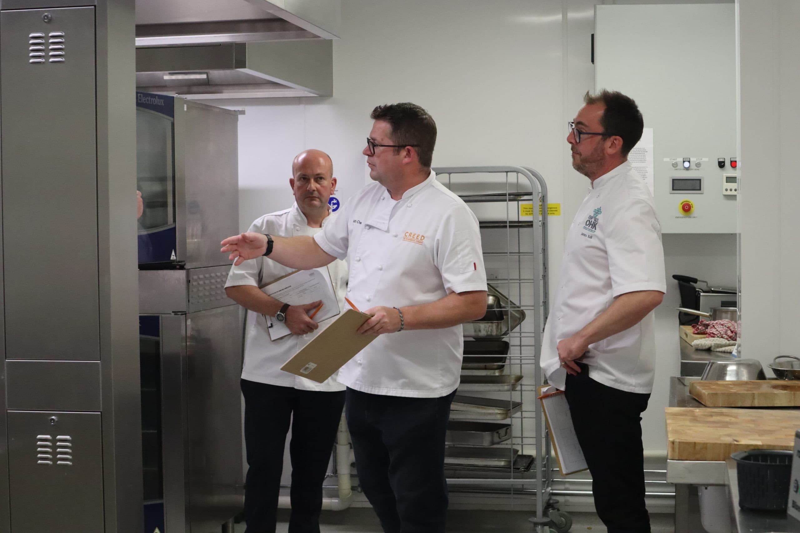 Three chefs in white uniforms discussing operations in a modern professional kitchen with stainless steel equipment