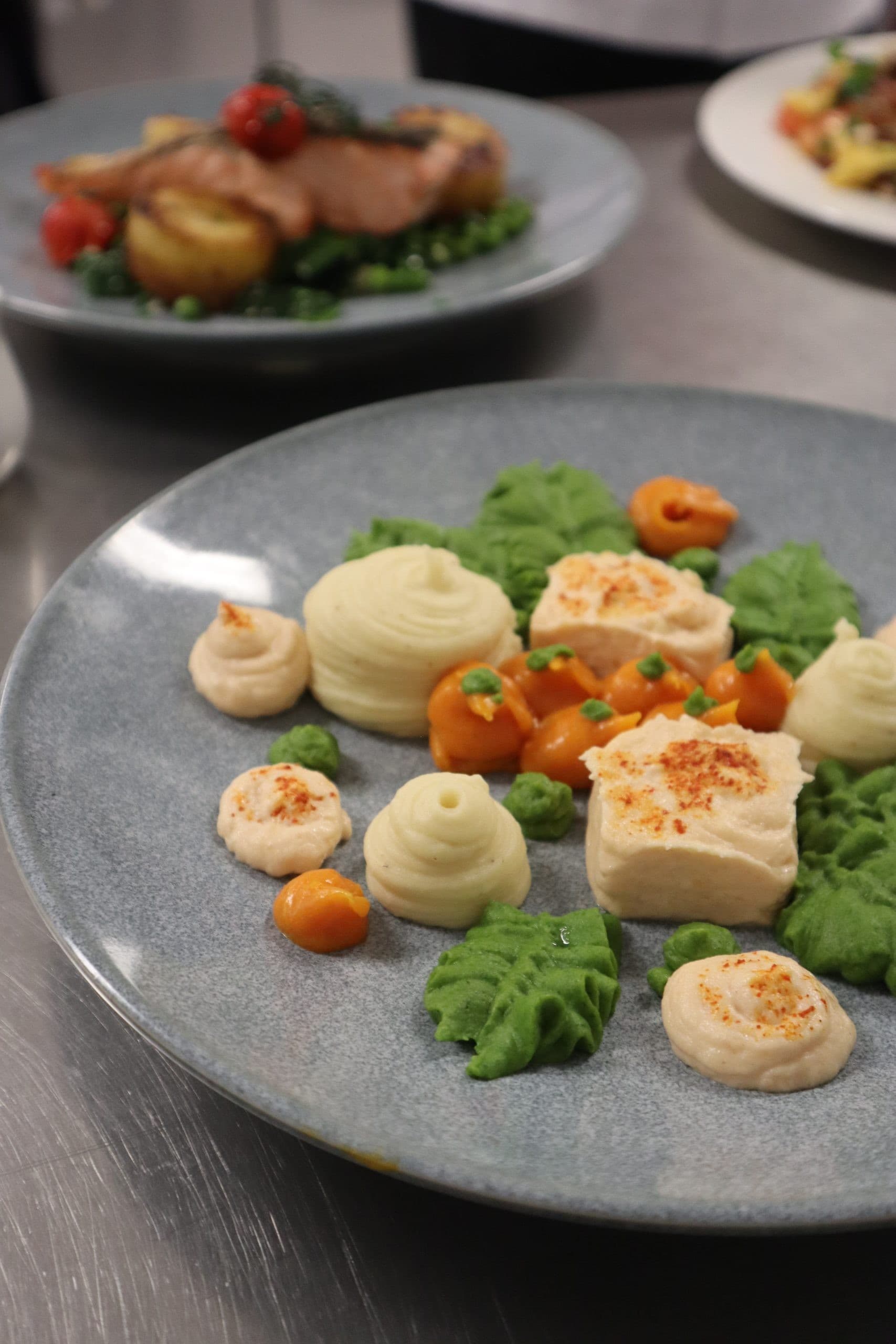 An elegantly plated dish featuring piped mashed potato swirls, roasted vegetables, and microgreens on a grey plate in a fine dining setting with additional plated dishes visible in the background.