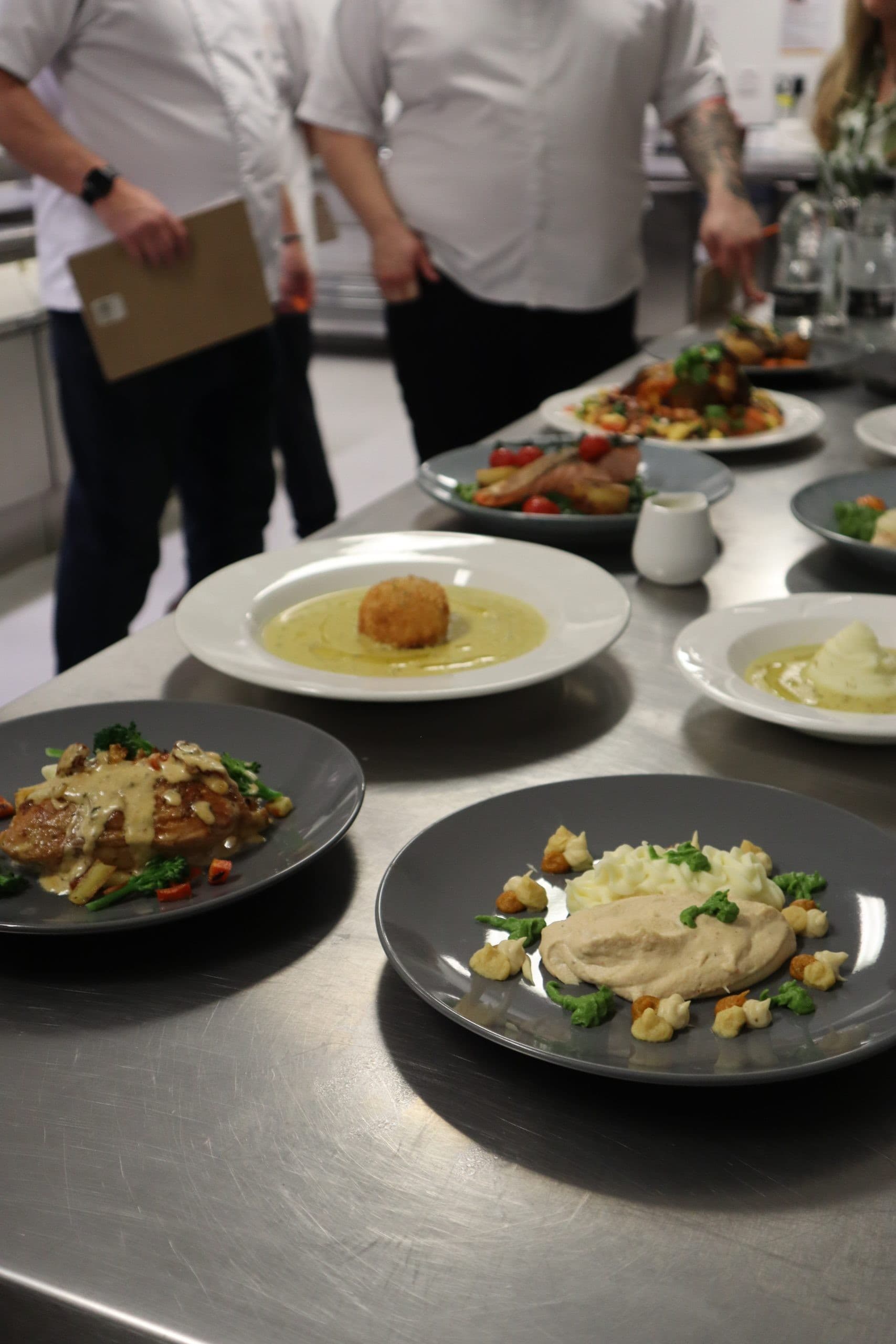 A culinary presentation of multiple gourmet dishes on white and grey plates arranged on a metal table, with chefs in white uniforms visible in the background of a professional kitchen setting.