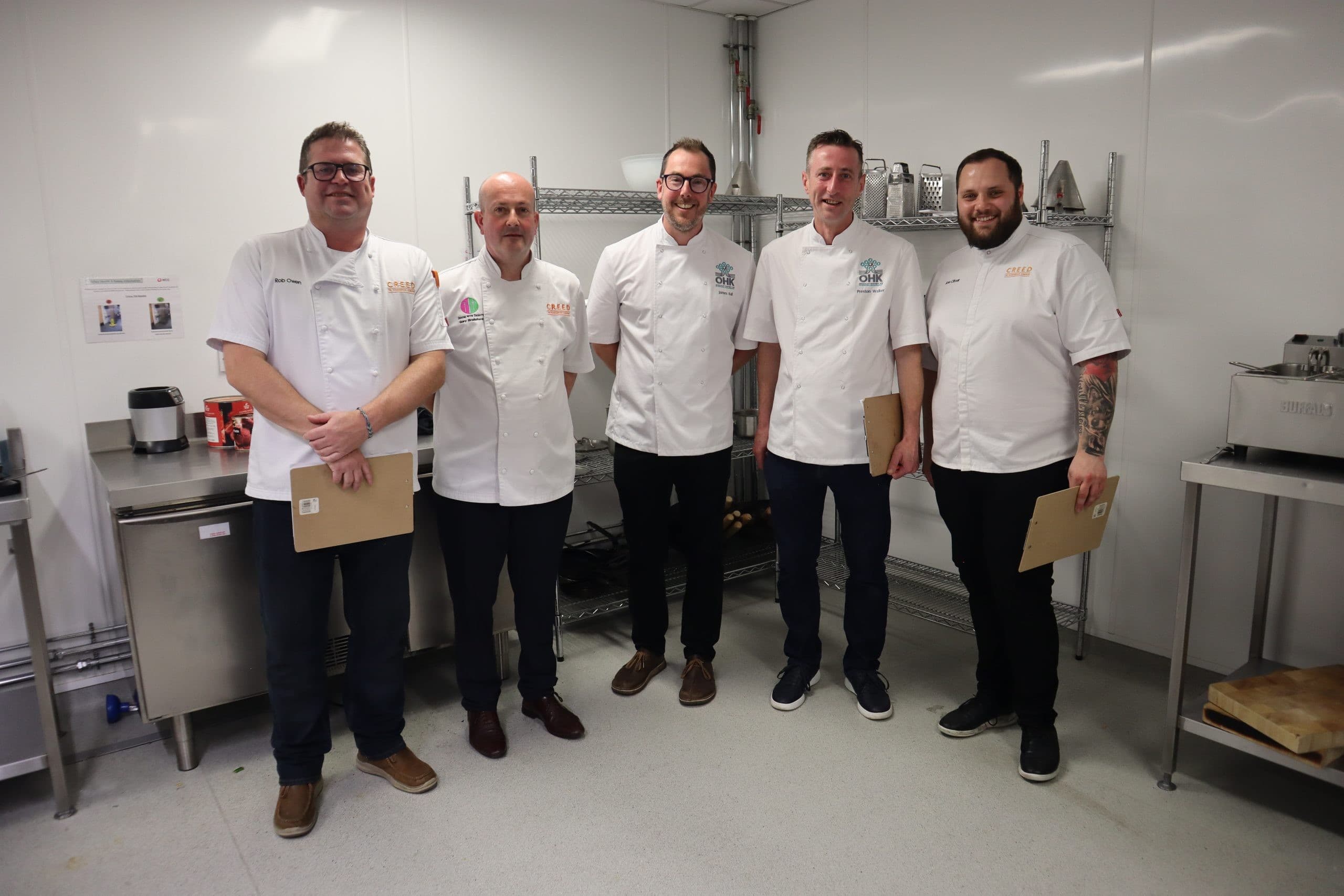 Five male chefs wearing white chef coats standing together in a professional kitchen holding cardboard boxes