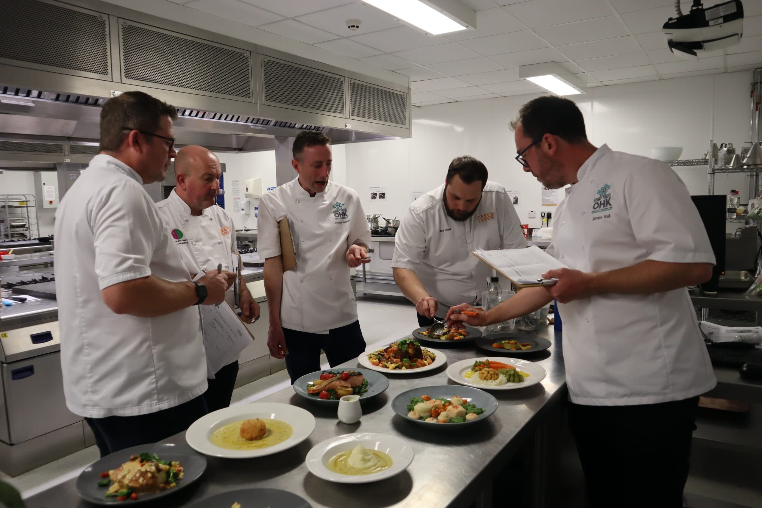 Five chefs in white chef coats gathered around a stainless steel counter examining multiple plated dishes in a professional kitchen setting