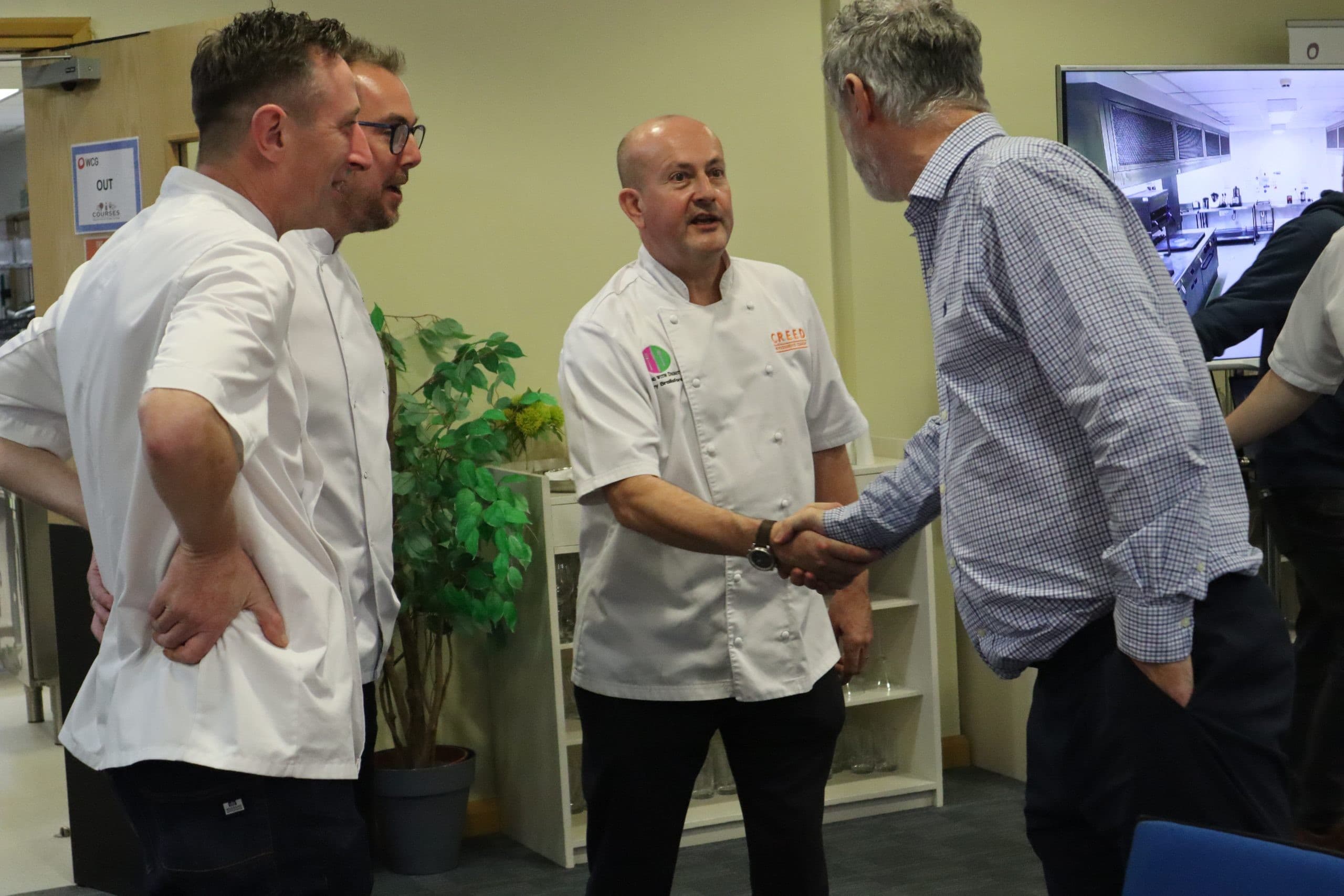 Three men in white chef coats shaking hands and conversing with a fourth man in a checkered shirt in a kitchen facility with yellow walls and a green potted plant.